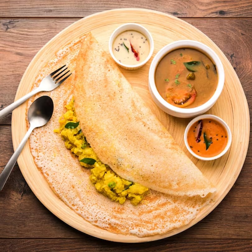 A golden-brown dosa with potato filling, served with sambar and two chutneys on a wooden plate.